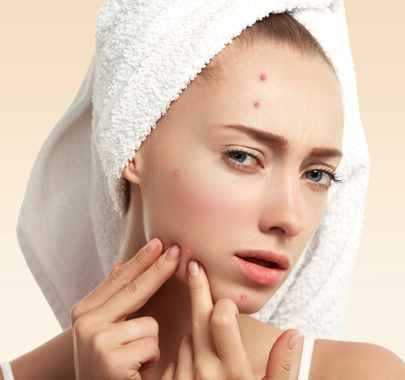 Close up isolated portrait of young woman squeezing pimple on her cheek in the bathroom. Cropped view of Caucasian female having skin problem, against blue wall background. Beauty and spa concept